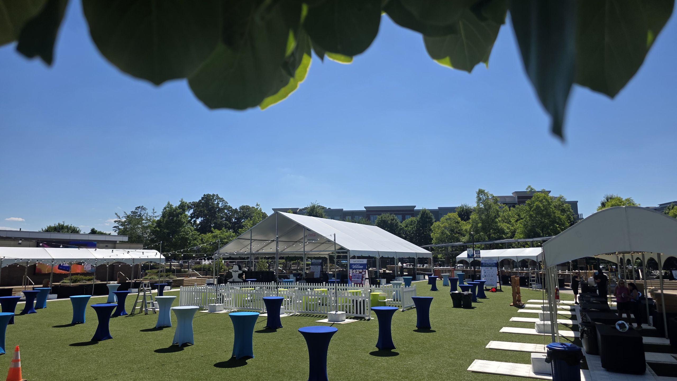 Outdoor event lawn with frame tents and blue-linen cocktail tables on a sunny day
