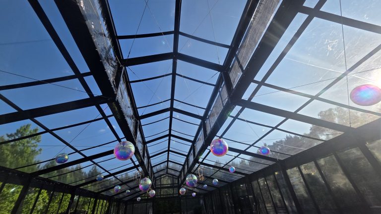 Looking up through the clear roof of a black-framed glass event structure with hanging decor