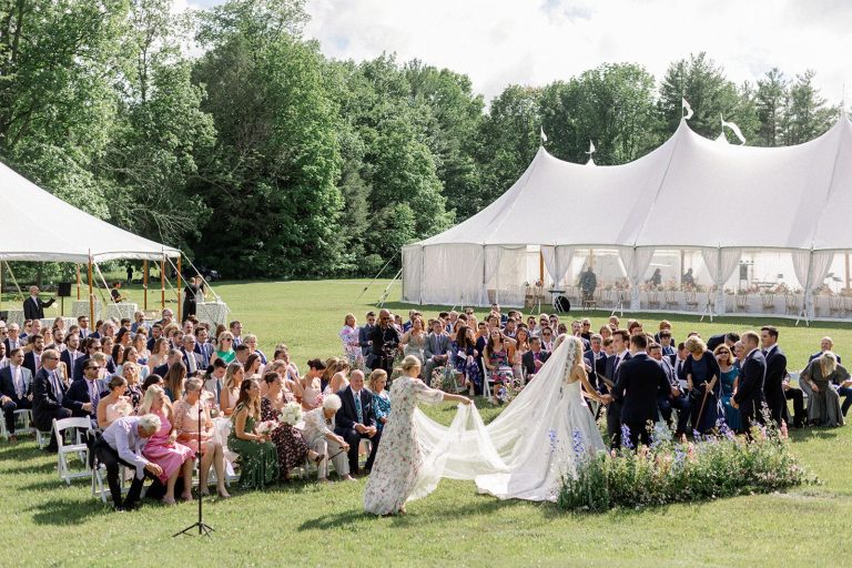 Outdoor wedding ceremony on a lawn with a white sailcloth tent behind the guests