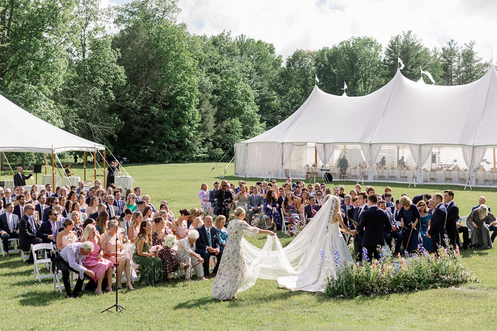 Outdoor wedding ceremony on a lawn with a white sailcloth tent behind the guests